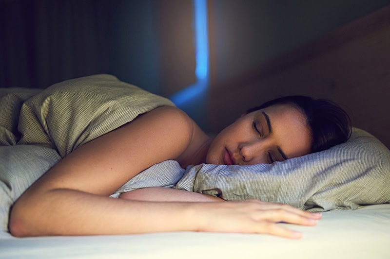 3 Health Benefits of Using Your AC While Sleeping. Photo of a young woman asleep in her bed.